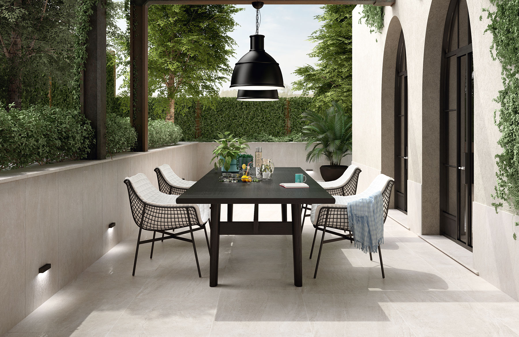 Modern outdoor dining area with a black table, wire chairs, and large light beige porcelain tiles, surrounded by greenery and arched doorways