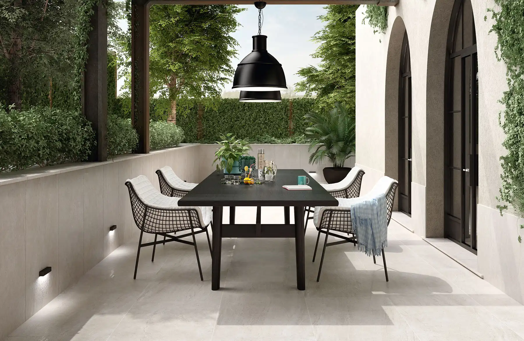 Modern outdoor dining area with a black table, wire chairs, and large light beige porcelain tiles, surrounded by greenery and arched doorways