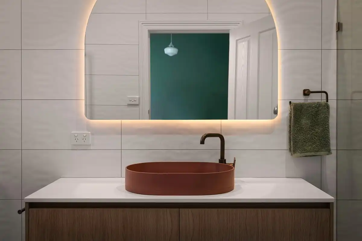 Bathroom vanity with a rust vessel sink, black faucet, arched backlit mirror, and white tiles with dark contrasting grout.