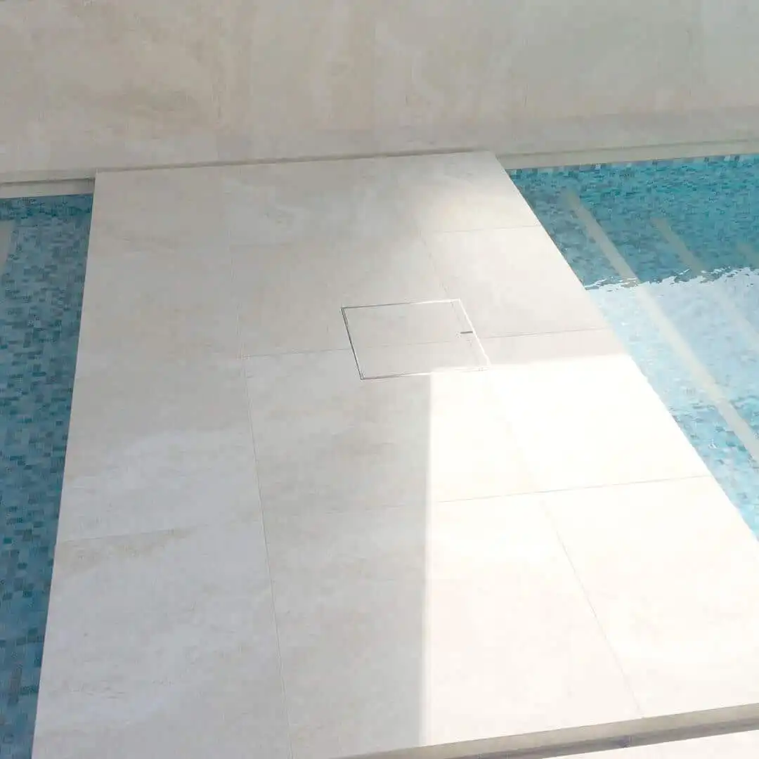 Light beige tiled pool deck with a concealed drain cover, surrounded by blue mosaic water, featuring neutral-colored grout.